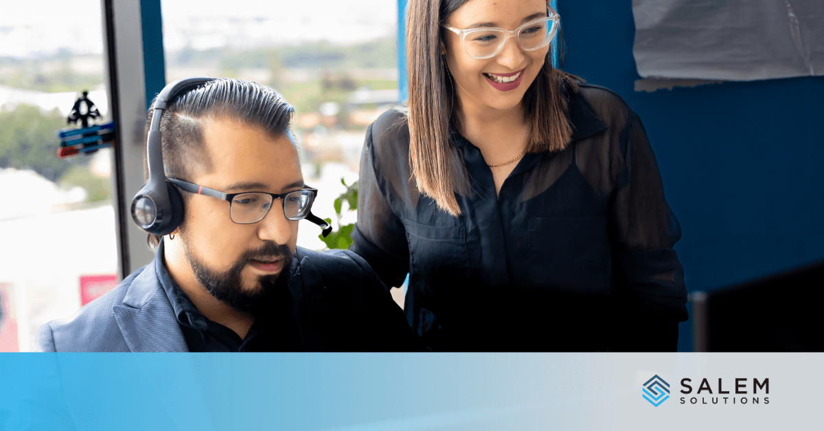 Two professionals wearing glasses, one seated and the other standing, are working together in an office.