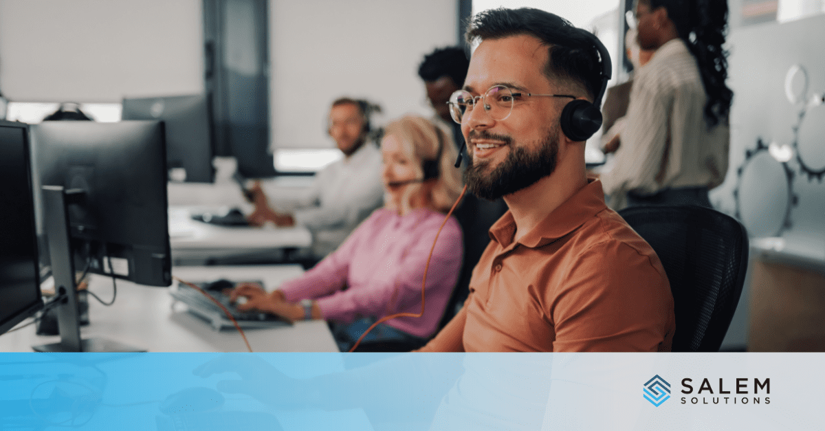 A man wearing a headset works at a computer in a modern office with other employees.