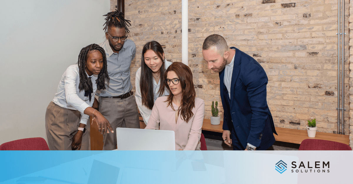 A diverse group of five professionals collaborates around a laptop in a modern office with a brick wall and wooden desk in the background, representing a high-performing customer service team.