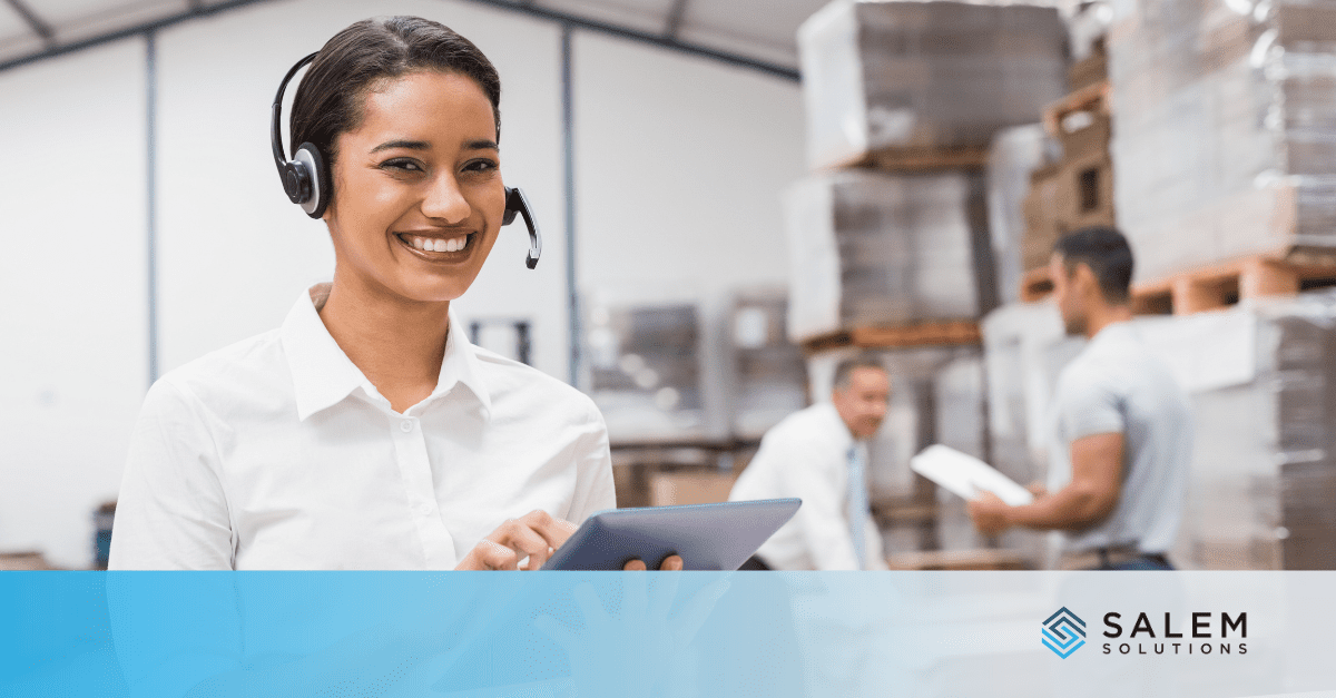 A retail call center agent in a white shirt uses a tablet and headset while coordinating operations near stacked pallets, illustrating effective holiday call center staffing.